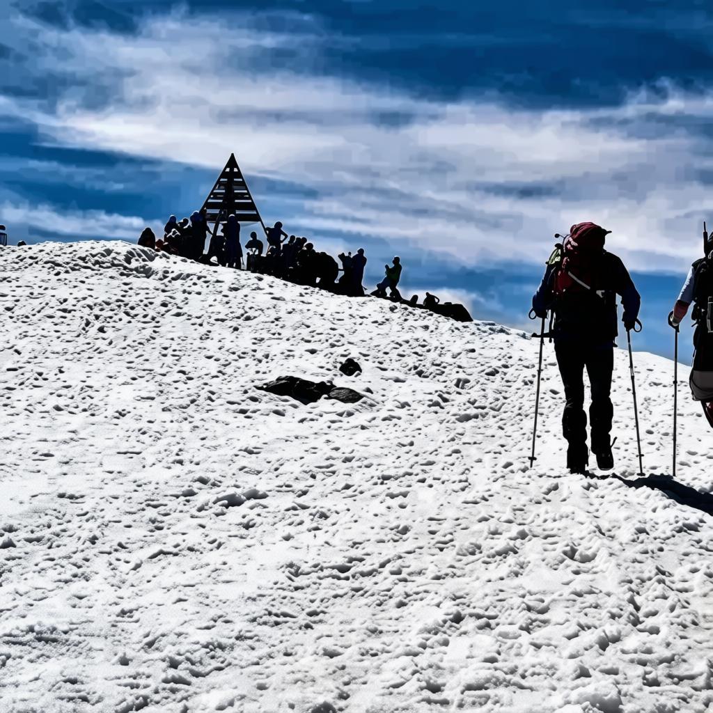 Trekking aventura en Marruecos: grupo de senderistas posando frente a aldea tradicional del Alto Atlas junto a paisaje montañoso nevado con estructura piramidal en la cima